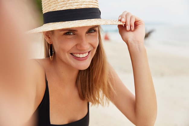 Colpo di donna sorridente dall'aspetto piacevole con cappello di paglia, ha un sorriso splendente, posa per selfie sullo sfondo dell'oceano, essendo di alto spirito mentre trascorre le vacanze estive in un luogo paradisiaco con l'amante