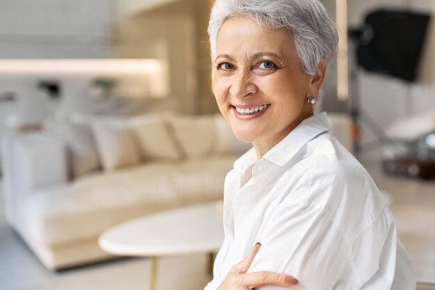 Colpo di felice cinquantenne donna in pensione con lentiggini e capelli grigi in posa su interni eleganti sfondo, indossa una camicia bianca, sorridente largamente davanti