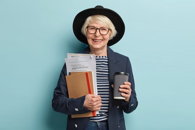 Colpo di felice donna d'affari con esperienza senior detiene documenti e portafogli, beve caffè da asporto, felice di firmare un contratto di successo, indossa cappello e cappotto alla moda, posa al coperto. Vecchio lavoratore occupato