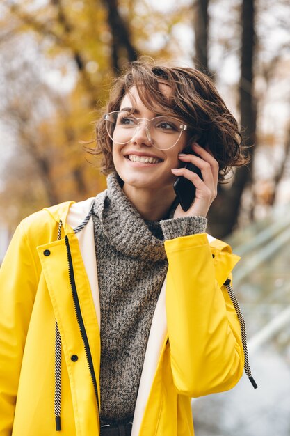 Colpo di giovane donna in giallo impermeabile e occhiali trascorrere del tempo fuori con piacevole conversazione mobile