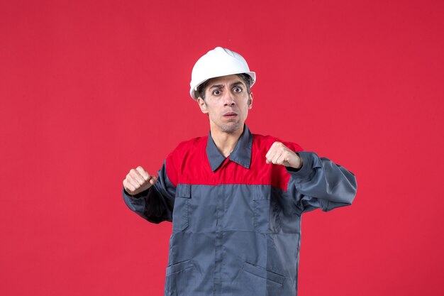 Colpo di mezzo corpo di giovane costruttore nervoso in uniforme con elmetto che stringe i pugni sul muro rosso isolato