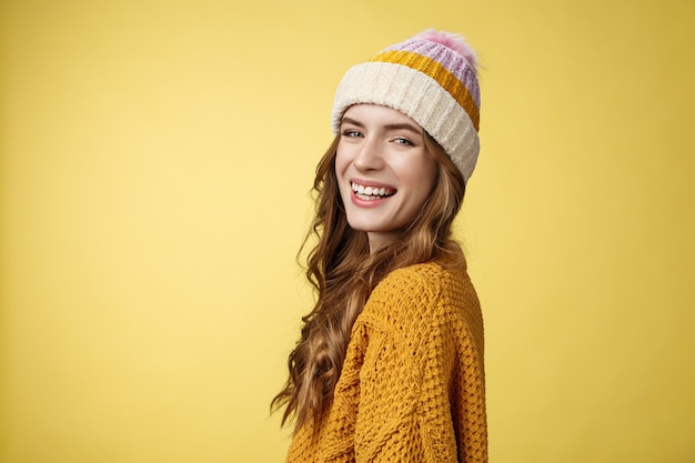 Colpo di profilo affascinante spensierata ragazza sorridente felice che gira la macchina fotografica sorridendo gioiosamente godendo le vacanze invernali divertendosi a sciare, in piedi divertito ridendo indossando un cappello maglione lavorato a maglia sfondo giallo