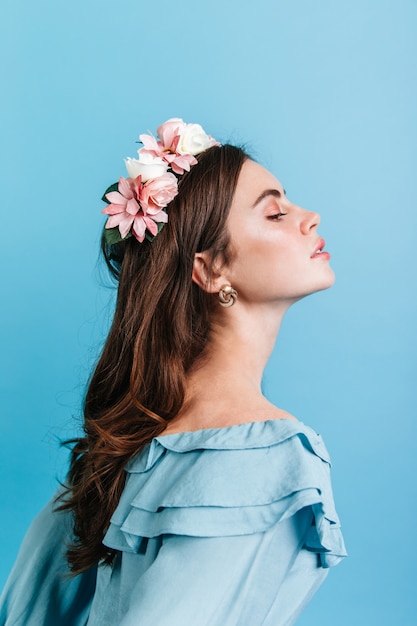 Colpo di profilo della ragazza aristocratica in camicetta con balza. Signora con fiori tra i capelli in posa con orgoglio contro il muro blu.