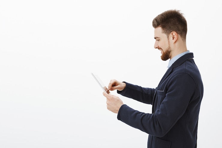 Foto gratuito colpo di profilo di determinato imprenditore elegante fiducioso e di successo con barba e acconciatura impressionante in abito elegante utilizzando tavoletta digitale sorridendo felice controllo reddito della società