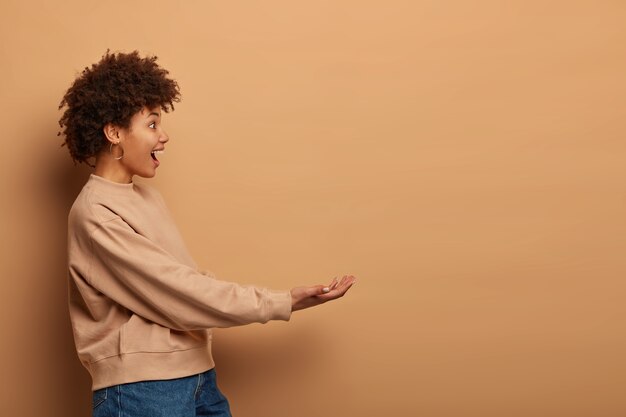 Colpo di profilo di donna afroamericana riccia felice finge di tenere qualcosa, tiene le mani tese, indossa jeans e maglione marrone sciolto, si leva in piedi contro il muro beige