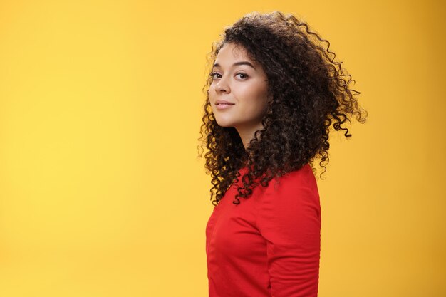 Colpo di profilo di donna europea 25s elegante e fresca attraente dai capelli ricci in abito rosso che gira alla macchina fotografica e sorride ampiamente con un'espressione spensierata felice, divertendosi su sfondo giallo.