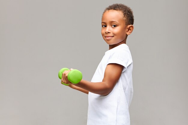 Colpo di profilo isolato del ragazzo africano gioioso bello in posa al muro bianco grigio utilizzando attrezzature per il fitness, tenendo il manubrio verde, costruzione muscolare del braccio, guardando con un sorriso felice