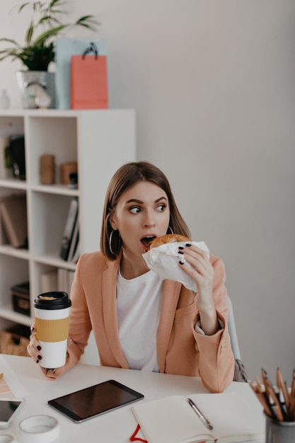 Colpo di signora sorpresa in giacca color pesca che mangia hamburger Donna d'affari che tiene un bicchiere di caffè sullo sfondo dell'ufficio