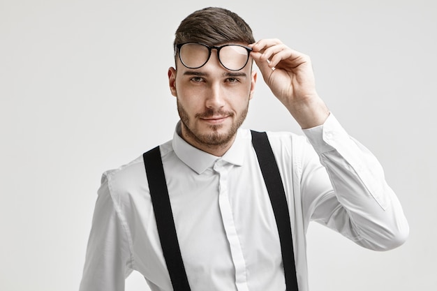 Colpo di studio isolato di attraente affascinante giovane barbuto impiegato in elegante camicia formale e bretelle sorridendo con gioia alla fotocamera, sollevando gli occhiali. Persone, stile di vita moderno e affari