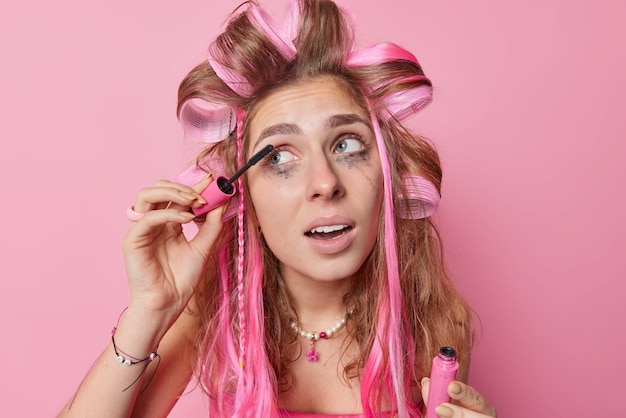 Colpo di testa di una giovane donna europea dai capelli lunghi applica i bigodini e il mascara distoglie lo sguardo si sottopone a trattamenti di bellezza si prepara per il primo appuntamento vuole apparire bella isolata su sfondo rosa