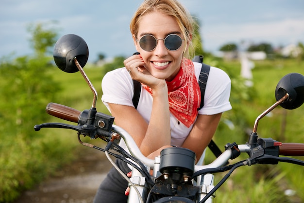 Colpo esterno del pilota femminile attivo positivo si siede su una moto veloce, indossa abiti alla moda, ha una pausa dopo la competizione di motociclisti in campagna. Persone, motociclismo e concetto di stile di vita