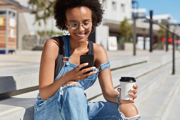 Colpo esterno di hipster felice che legge commenti sul sito web, si concentra sullo schermo del telefono cellulare, tiene un caffè da asporto, si siede sulle scale, indossa abiti in denim, gode di Internet ad alta velocità in roaming.