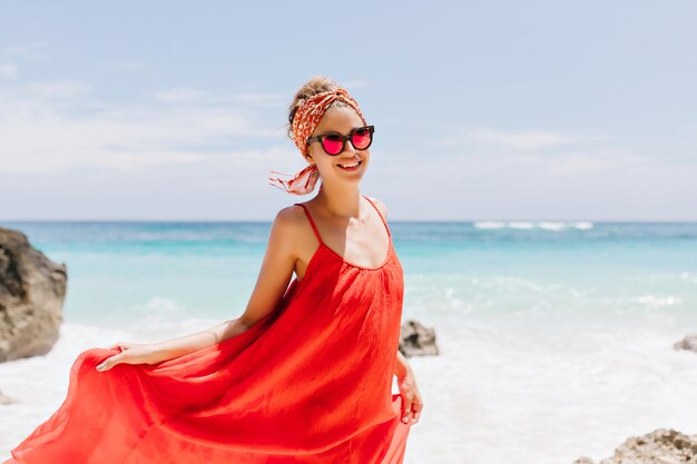 Colpo esterno di raffinata ragazza abbronzata in posa con piacere in spiaggia. Ritratto di splendida giovane signora che gioca con il vestito rosso e sorridente in spiaggia.