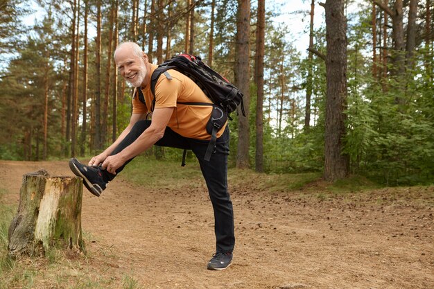 Colpo estivo all'aperto di maschio anziano in forma sana con zaino in posa nella foresta con il piede sul mozzo, allacciare i lacci delle scarpe da ginnastica, prepararsi per una lunga salita, fare escursioni con un sorriso felice