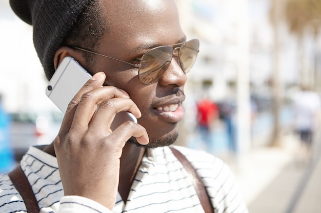 Colpo in testa della persona di colore in tonalità che hanno conversazione telefonica il giorno di molla soleggiato, godendo di bella passeggiata sulle vie della località turistica. Persone in vacanza. Giovani e viaggi. Umano e tecnologia