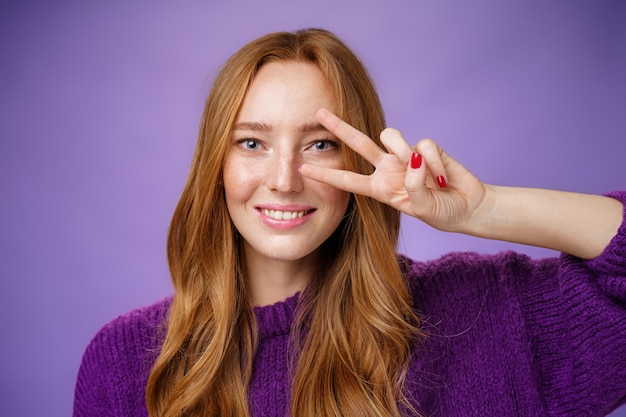 Colpo in testa di studentessa rossa felice amichevole e ottimista in maglione viola che mostra vittoria o gesto di pace sopra l'occhio come mossa di ballo della discoteca che sorride ampiamente sopra fondo viola.