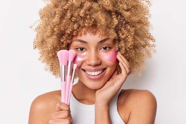 Colpo in testa di una donna positiva con capelli ricci e folti tocca il viso sorride dolcemente applica cerotti idrogel rosa sotto gli occhi usa pennelli cosmetici per applicare il trucco isolato su un muro bianco