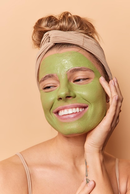 Colpo in testa di una giovane donna piuttosto soddisfatta tiene le mani sul viso guarda in basso sorride applica a denti stretti la maschera nutriente verde ha i capelli pettinati indossa la fascia pone le spalle nude contro il muro marrone dello studio