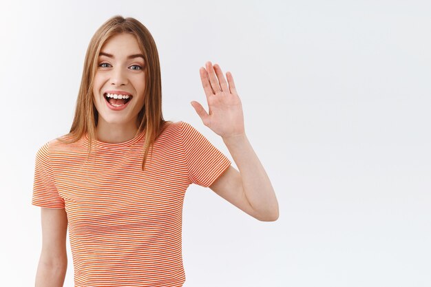 Colpo in vita amichevole, bella giovane donna in t-shirt a righe, alzando la mano e salutandoti, adorabile sorridente piacevolmente saluta, ciao, in piedi sfondo bianco accogliendo il cliente in negozio