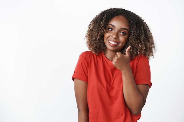 Colpo in vita di simpatica donna afroamericana di supporto che mostra pollice in su in modo simile e approvazione inclinando la testa e sorridendo ampiamente per allietare l'amico e incoraggiare gli sforzi eccellenti sul muro bianco