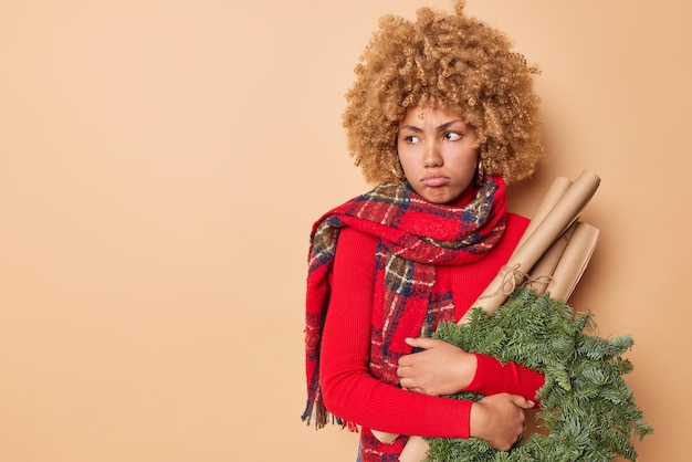 Colpo in vita di una donna cupa sconvolta ha un cattivo umore indossa un maglione e una sciarpa porta una corona di abete verde e i rulli di carta distoglie lo sguardo si sente dispiaciuto isolato su sfondo beige copia spazio lontano