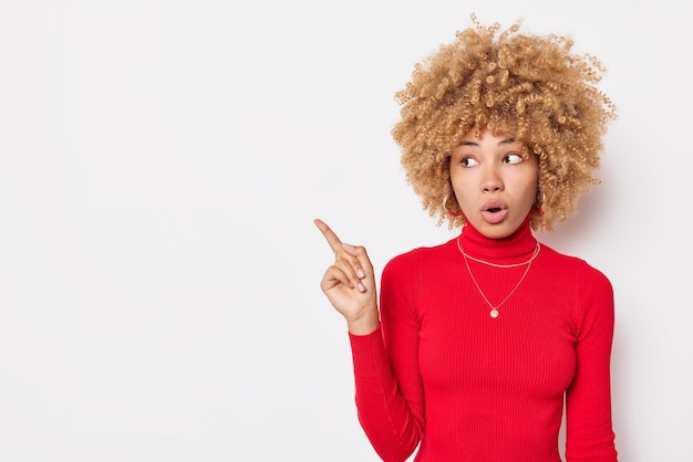 Colpo in vita di una donna stupita con i capelli ricci che punta in uno spazio vuoto si sente scioccata indossa un dolcevita rosso isolato su sfondo bianco La donna curiosa fa domande sulle pose del prodotto all'interno