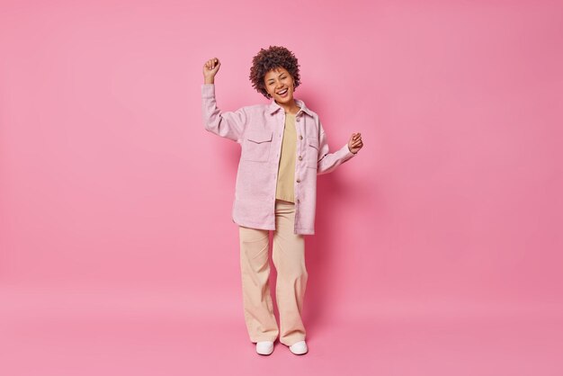 Colpo integrale della donna dai capelli ricci alla moda si diverte a ballare spensierato tiene le braccia alzate con eleganti pantaloni camicia e scarpe da ginnastica posa contro il muro rosa