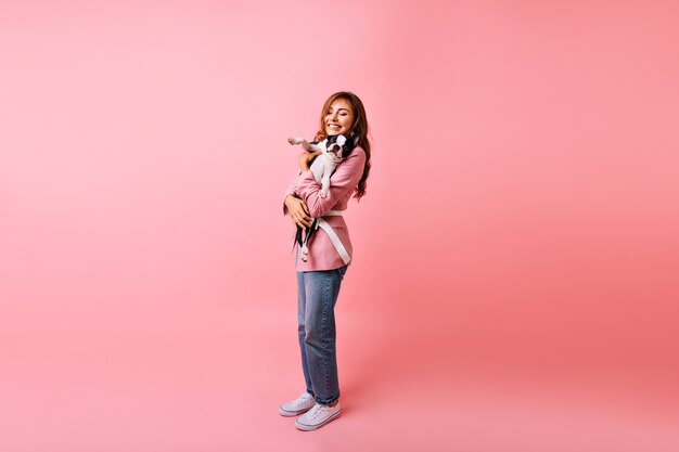 Colpo integrale della ragazza adorabile in jeans alla moda che tengono il bulldog francese. Ritratto dell'interno della donna dai capelli lunghi felice in posa con il suo animale domestico.