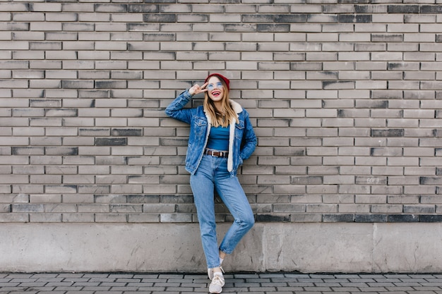 Colpo integrale della signora emotiva in jeans in piedi con il segno di pace sul muro di mattoni. Donna abbastanza formosa in abbigliamento denim in posa sulla strada vicino al muro.