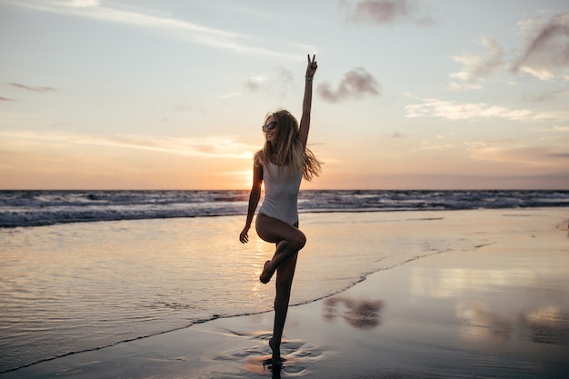 Colpo integrale di adorabile ragazza sottile in piedi su una gamba sola sulla costa dell'oceano.