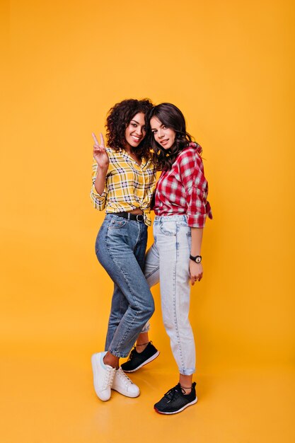 Colpo integrale di amiche alla moda in jeans di mamme. Ragazze con capelli ricci scuri in posa di buon umore.