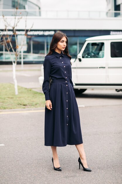 Colpo integrale di giovane signora abbastanza bella in abito nero elegante e décolleté nere su sfondo di città. Stile e concetto di moda