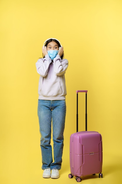 Colpo integrale di turista asiatico sorridente felice, ragazza in cuffia, ascoltando musica in aeroporto, indossando maschera medica, in piedi con la valigia, sfondo giallo
