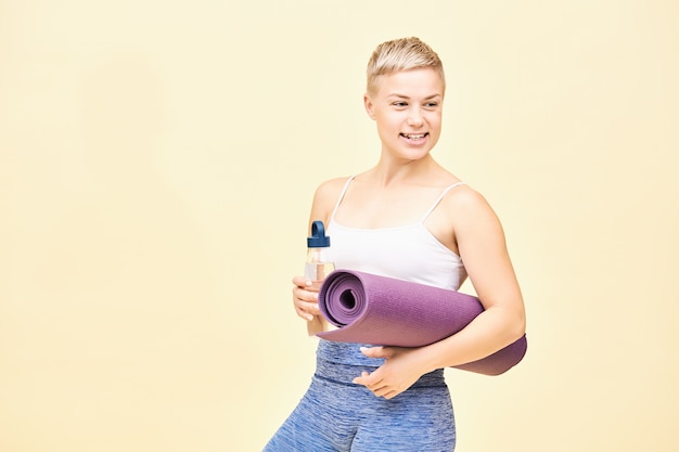 Colpo isolato di bella felice giovane istruttrice femminile con capelli tinti di folletto andando per allenamento portando una bottiglia di vetro non usa e getta con acqua e stuoia di yoga arrotolata, distogliendo lo sguardo con un ampio sorriso