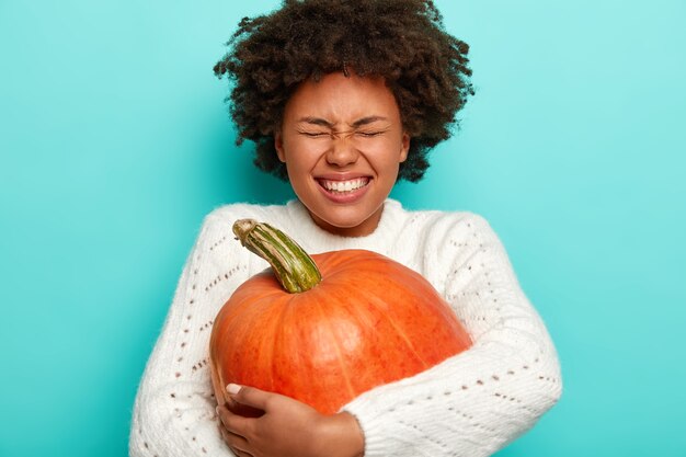 Colpo isolato di donna ipermotiva gioiosa con capelli ricci afro, raccoglie grande zucca, indossa un maglione caldo, isolato su blu.