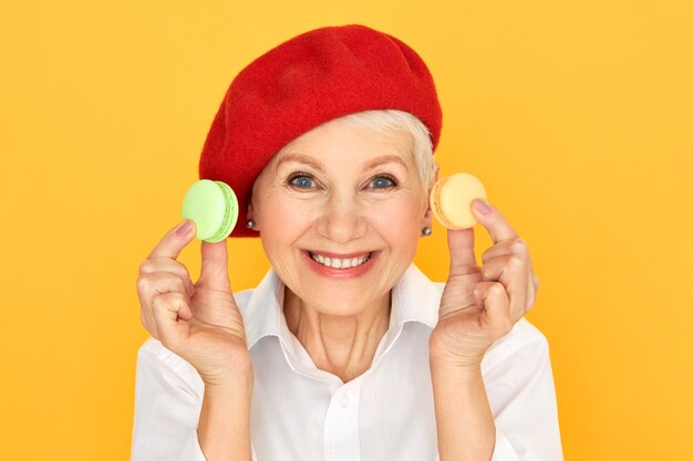 Colpo isolato di pensionato femmina senior divertente adorabile nel cofano rosso che sorride ampiamente alla macchina fotografica che tiene i biscotti francesi variopinti.