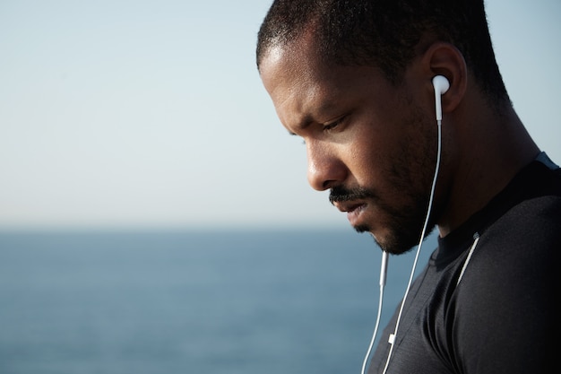 Colpo laterale dell'uomo triste dell'afroamericano che osserva giù e che ascolta la musica malinconica in cuffie con il fronte serio e pensieroso.