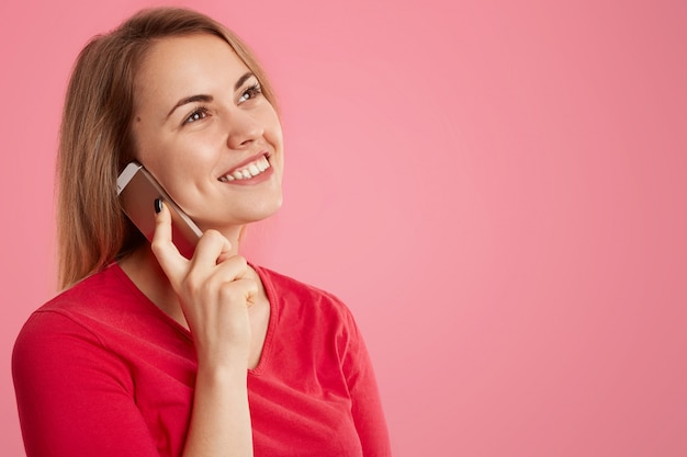 Colpo laterale di attraente donna felice ha un'espressione sognante, parla al cellulare, gode di una conversazione con un amico, indossa un maglione rosso