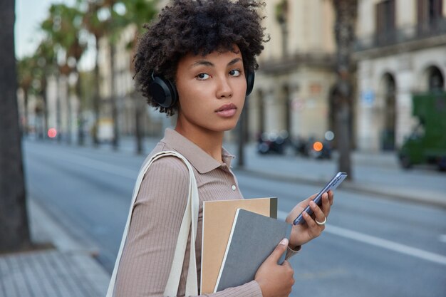 Colpo laterale di studentessa premurosa usa smartwatch e cuffie ascolta webinar educativo tramite cuffie mentre va all'università tiene quaderni distoglie lo sguardo con espressione pensierosa