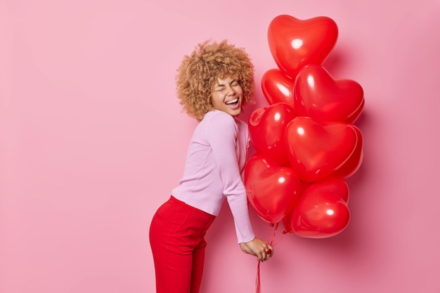 Colpo laterale di una donna dai capelli ricci felicissima che indossa un maglione casual e pantaloni rossi tiene un mazzo di palloncini gonfiati a cuore si diverte durante la celebrazione isolato su sfondo rosa Concetto di vacanze