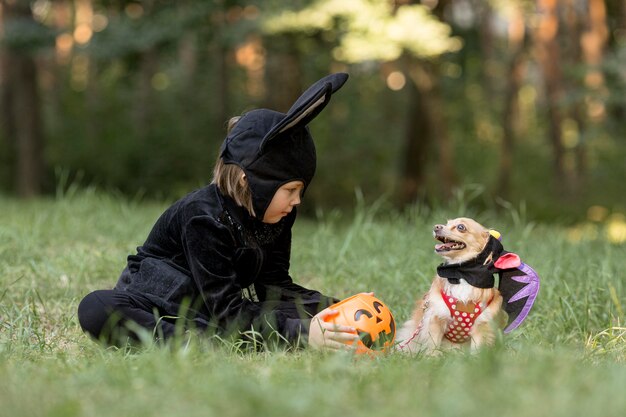 Colpo lungo del ragazzino in costume da pipistrello e cane