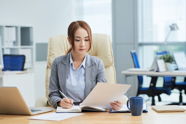 Colpo medio di giovane donna asiatica in vestito che si siede allo scrittorio in ufficio e che legge documento