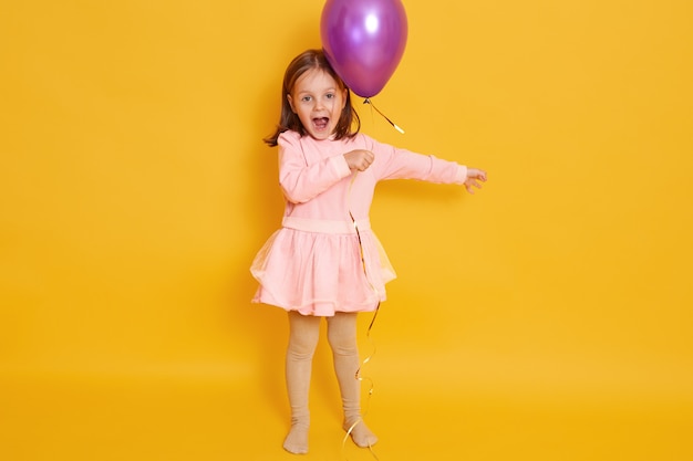 Colpo orizzontale della bambina con l'aerostato viola isolato sopra giallo tutto il bambino femminile che grida qualcosa, celebrando il herbirtday, bambino che porta il vestito roseo e che ha capelli scuri.