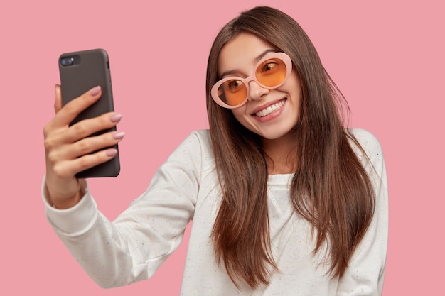 Colpo orizzontale della bella ragazza dai capelli scuri sorride felicemente, inclina la testa, tiene il cellulare, registra vlog, prende selfie, indossa occhiali da sole alla moda e maglione casual bianco, isolato sopra il muro rosa