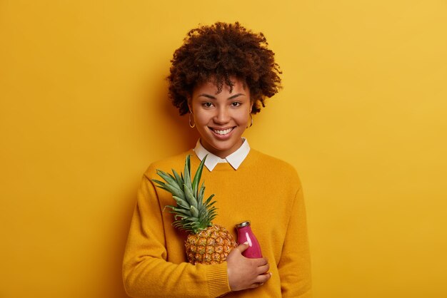 Colpo orizzontale della ragazza felice dall'aspetto piacevole con l'acconciatura afro, tiene ananas maturo e frullato, posa con frutta esotica, ha un ampio sorriso a trentadue denti, sguardo diretto, isolato sulla parete gialla