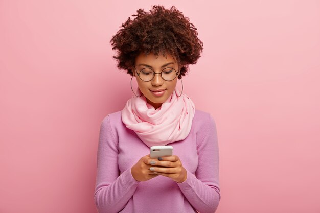 Colpo orizzontale di bella giovane donna con acconciatura afro, utilizza il dispositivo smartphone, concentrato sul display, invia messaggi di testo, connesso a Internet wireless indossa sciarpa viola intorno al collo