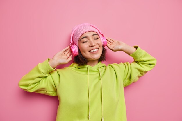 Colpo orizzontale di felice adolescente asiatica gode di una buona qualità del suono in nuove cuffie ascolta la musica preferita chiude gli occhi dai sorrisi di soddisfazione indossa ampiamente felpa con cappuccio e cappello isolato sul muro rosa