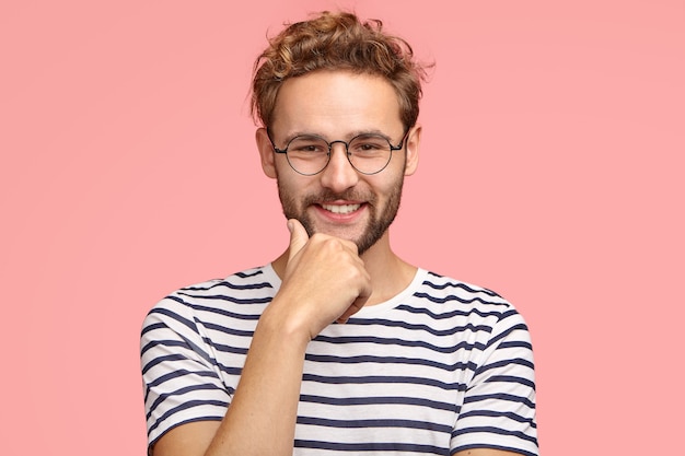 Colpo orizzontale di ragazzo soddisfatto di sé sicuro con un sorriso piacevole, tiene la mano sul mento