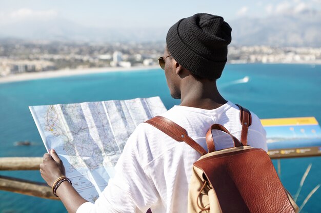 Colpo posteriore all'aperto del turista dalla pelle scura con zaino in pelle sulle spalle con guida della carta in mano, di fronte a una splendida vista meravigliosa della costa del mare europeo, in piedi sulla piattaforma turistica