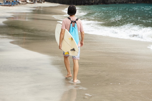 Colpo posteriore del surfista alla moda con la borsa blu che trasporta la sua tavola da surf bianca
